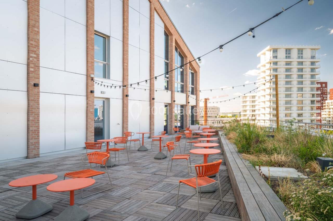Terrasse aménagée avec tables et chaises orange, entourée de plantes et éclairée par des guirlandes lumineuses.