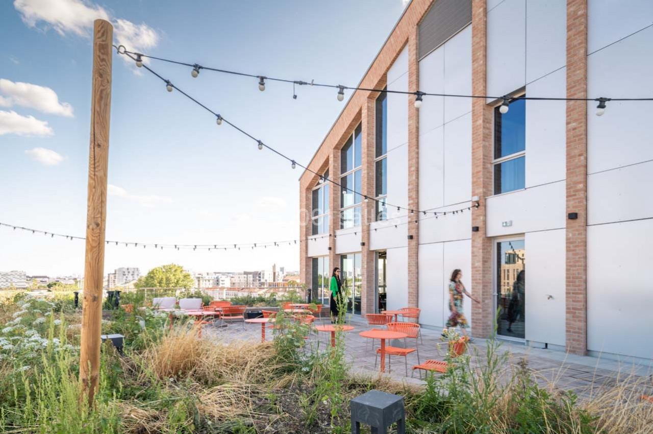 Terrasse aménagée avec des tables et chaises orange, entourée de végétation, près d'un bâtiment moderne en briques.