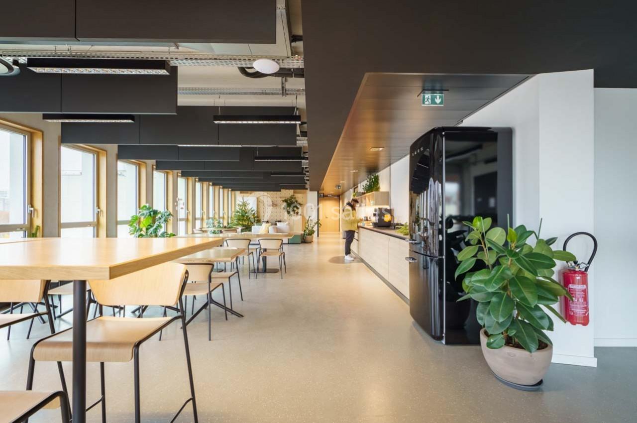 Espace de coworking lumineux avec tables, chaises, plantes, cuisine équipée et grandes fenêtres.