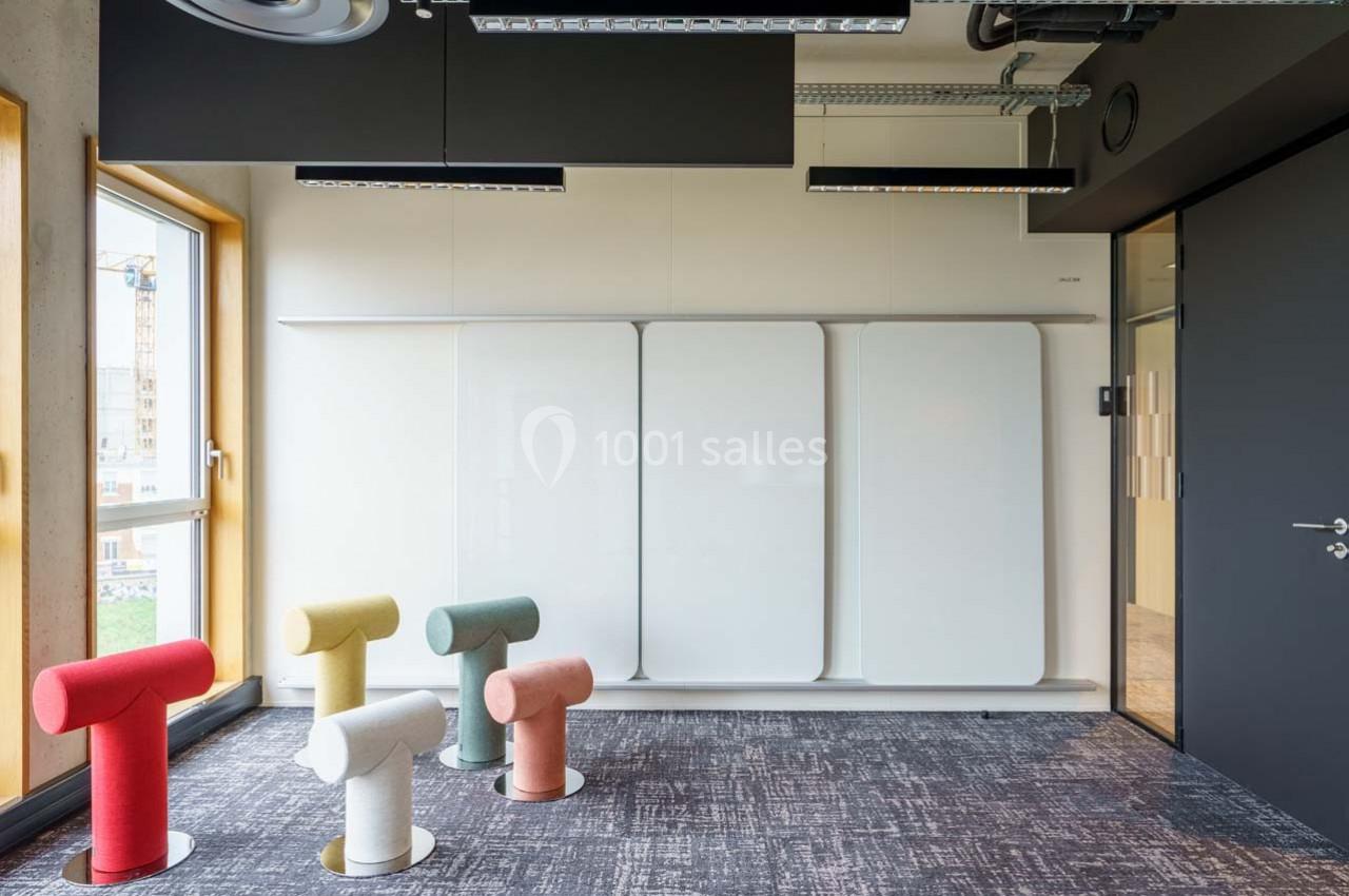 Salle de réunion moderne avec tableaux blancs muraux, sièges colorés en forme de T et grandes fenêtres.
