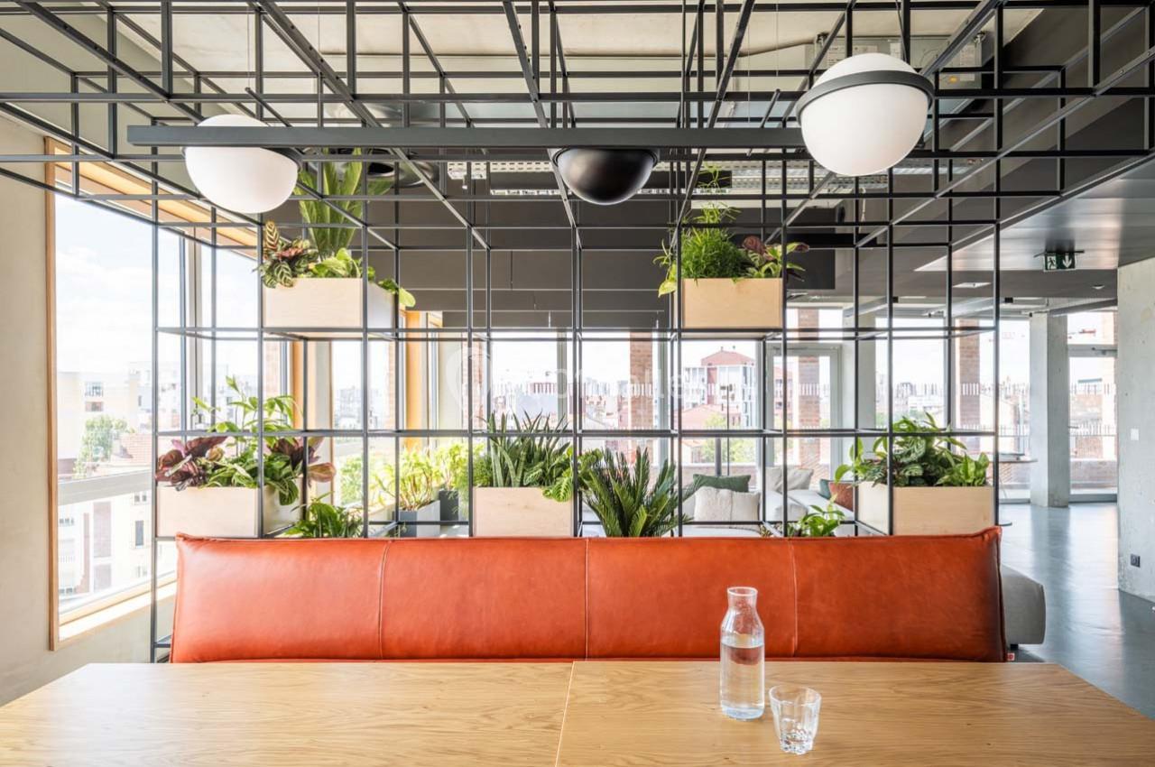 Espace lumineux avec cloison en métal ornée de plantes, table en bois, banquette en cuir orange et vue sur la ville.