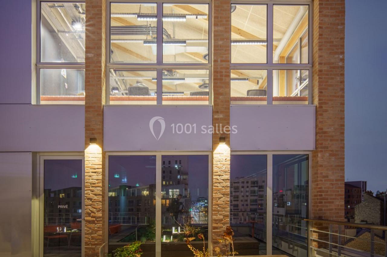 Façade d'un bâtiment moderne avec grandes baies vitrées illuminées, laissant entrevoir un intérieur aménagé.