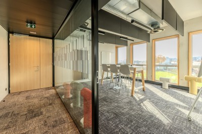 Salle de réunion moderne avec deux grandes tables en bois, chaises orange et baies vitrées donnant sur des bâtiments urbains.