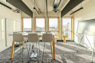 Salle de réunion moderne avec deux grandes tables en bois, chaises orange et baies vitrées donnant sur des bâtiments urbains.