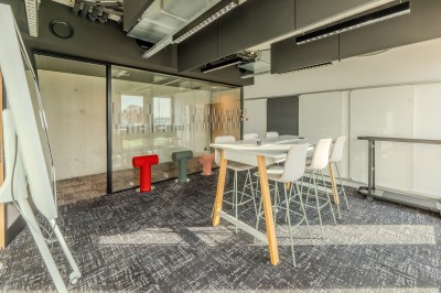 Salle de réunion moderne avec deux grandes tables en bois, chaises orange et baies vitrées donnant sur des bâtiments urbains.