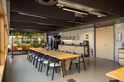 Salle de réunion moderne avec deux grandes tables en bois, chaises orange et baies vitrées donnant sur des bâtiments urbains.