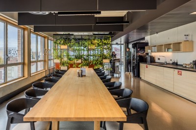 Salle de réunion moderne avec deux grandes tables en bois, chaises orange et baies vitrées donnant sur des bâtiments urbains.