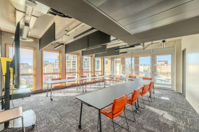 Salle de réunion moderne avec deux grandes tables en bois, chaises orange et baies vitrées donnant sur des bâtiments urbains.