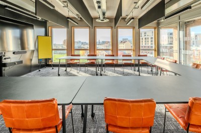 Salle de réunion moderne avec deux grandes tables en bois, chaises orange et baies vitrées donnant sur des bâtiments urbains.