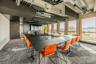 Salle de réunion moderne avec deux grandes tables en bois, chaises orange et baies vitrées donnant sur des bâtiments urbains.