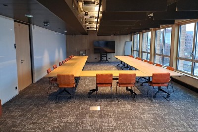 Salle de réunion moderne avec deux grandes tables en bois, chaises orange et baies vitrées donnant sur des bâtiments urbains.