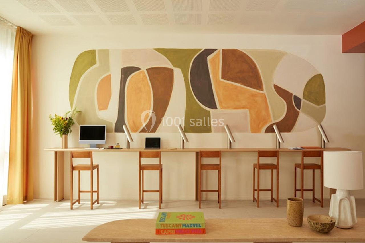 Espace de travail lumineux avec un long bureau, des chaises en bois et une fresque murale abstraite aux tons chauds.