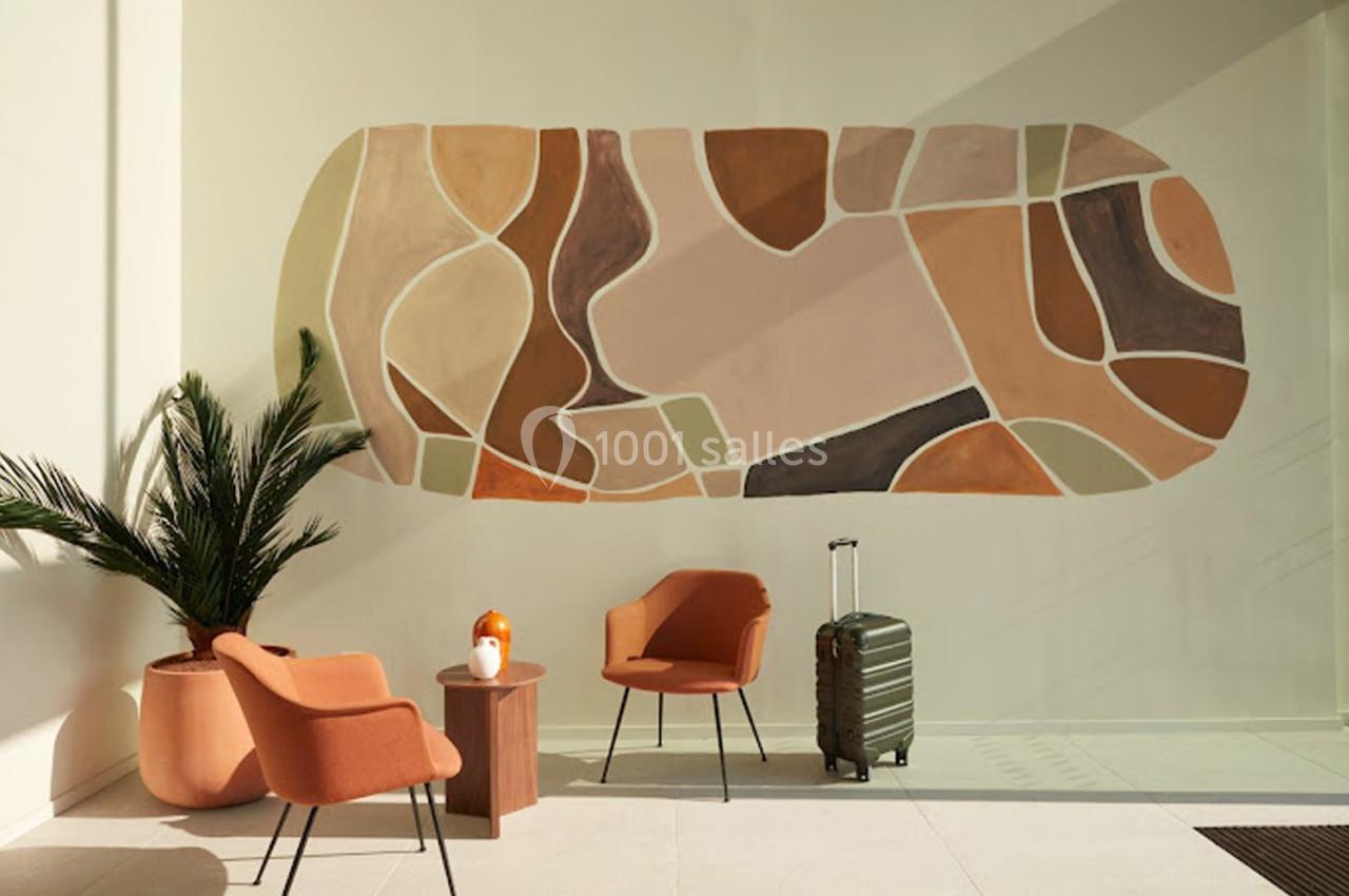 Mur décoré d'une fresque abstraite dans des tons terre, avec fauteuils orange, plante verte et valise posée au sol.