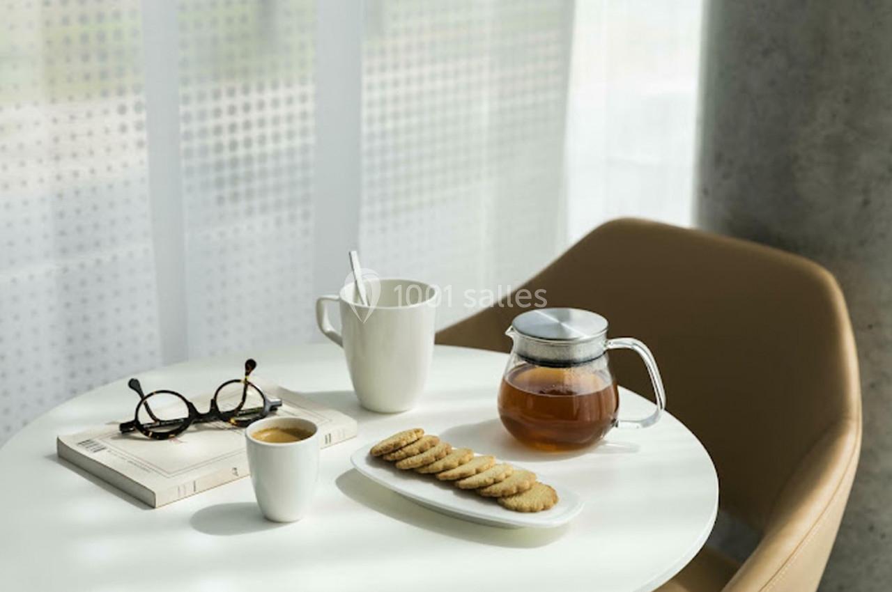 Table ronde avec une théière, une tasse, des biscuits, un café, des lunettes et un livre près d'une fenêtre lumineuse.