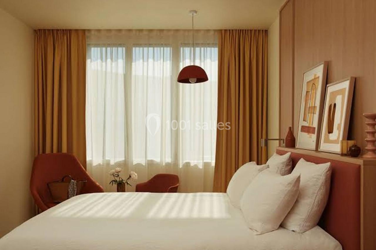 Chambre lumineuse avec lit double, fauteuils rouges, rideaux ocres et décorations murales dans un style moderne.