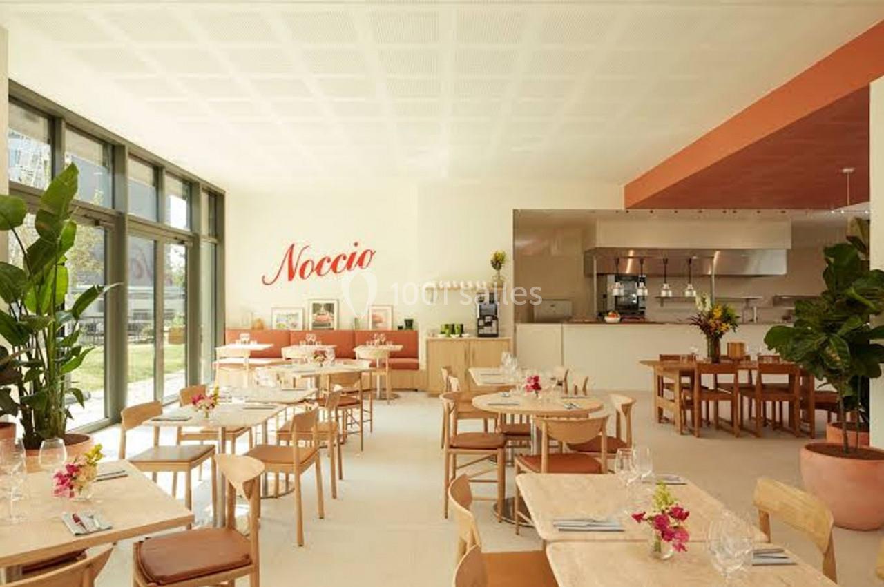 Salle lumineuse d'un restaurant moderne avec des tables en bois, des plantes et une cuisine ouverte en arrière-plan.