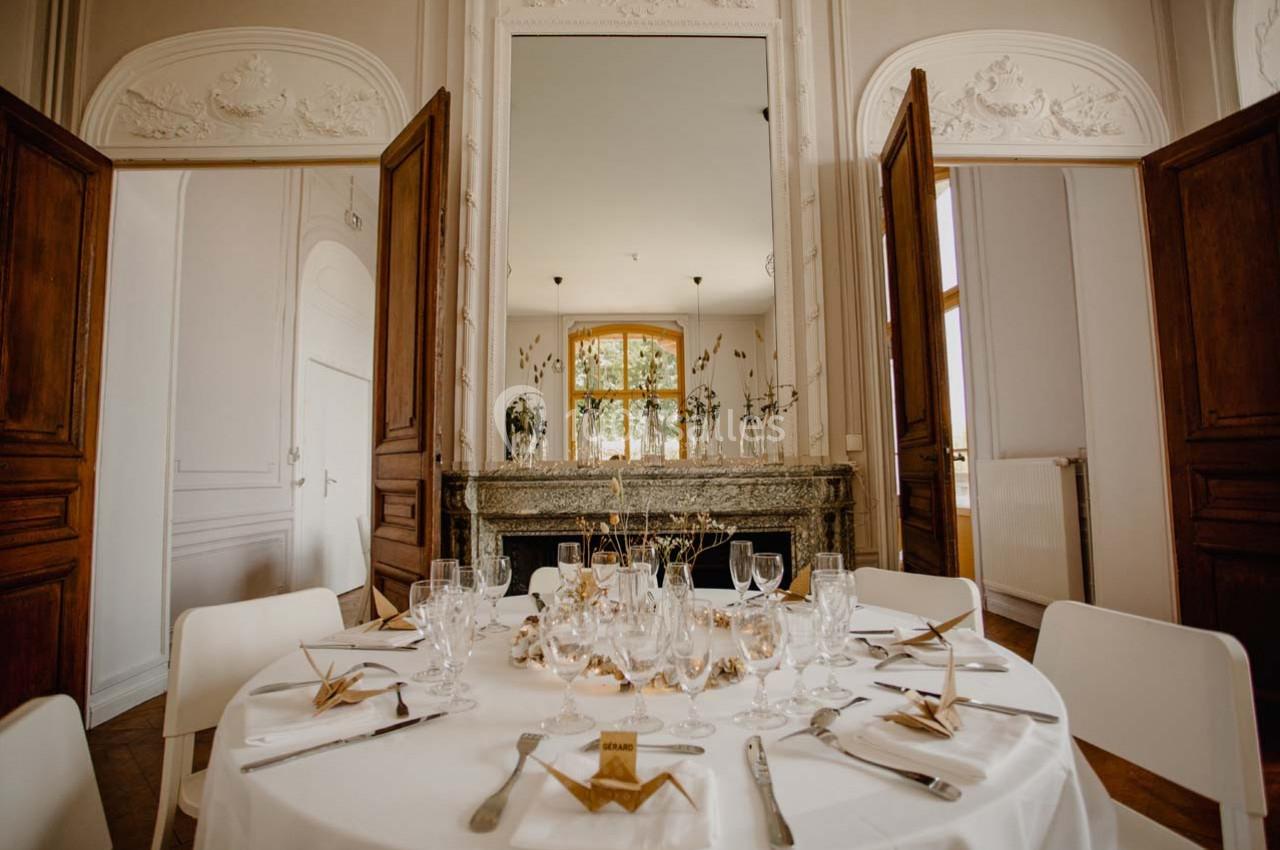 Table ronde dressée avec vaisselle élégante dans une salle lumineuse aux murs ornés et portes en bois ouvertes.