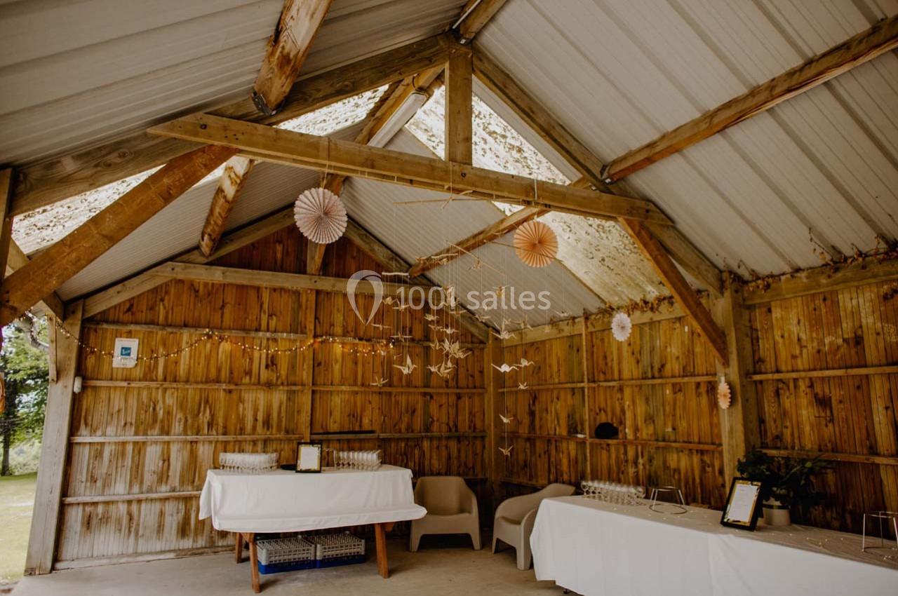 Intérieur d'une grange en bois décorée pour un événement, avec guirlandes lumineuses, tables et origamis suspendus.