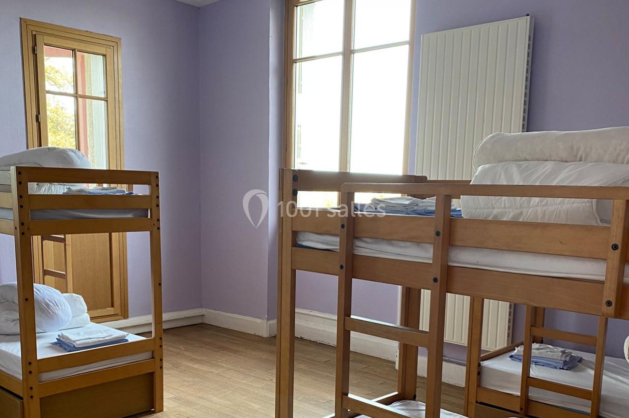 Chambre avec lits superposés en bois, draps pliés, parquet au sol, murs violets et grande fenêtre laissant entrer la lumière.