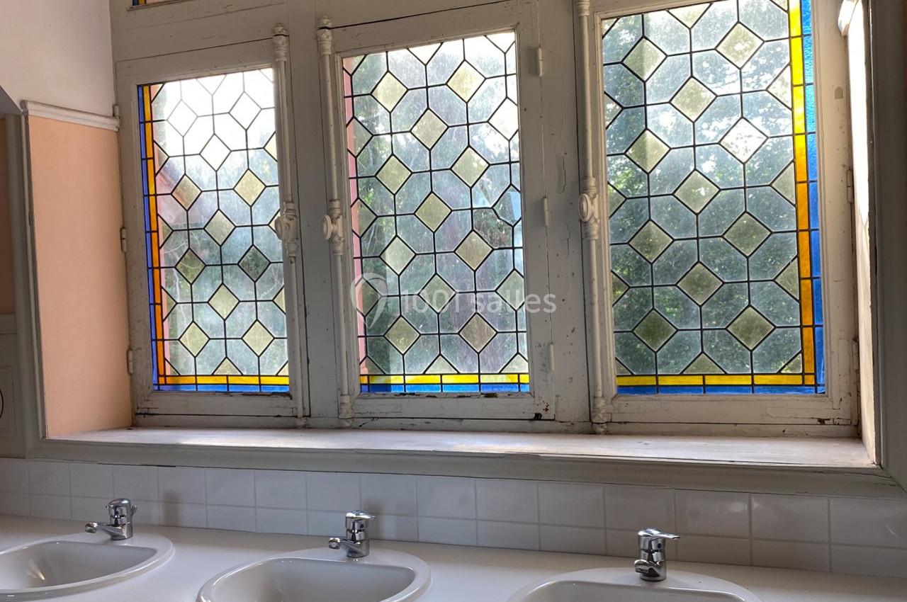 Fenêtres à vitraux colorés éclairant une salle avec trois lavabos blancs alignés sur un plan de travail carrelé.