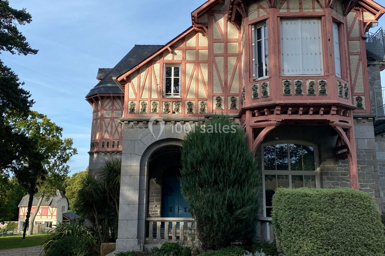 Façade d'une maison à colombages avec des détails en bois rouge, entourée de végétation et éclairée par la lumière du jour.