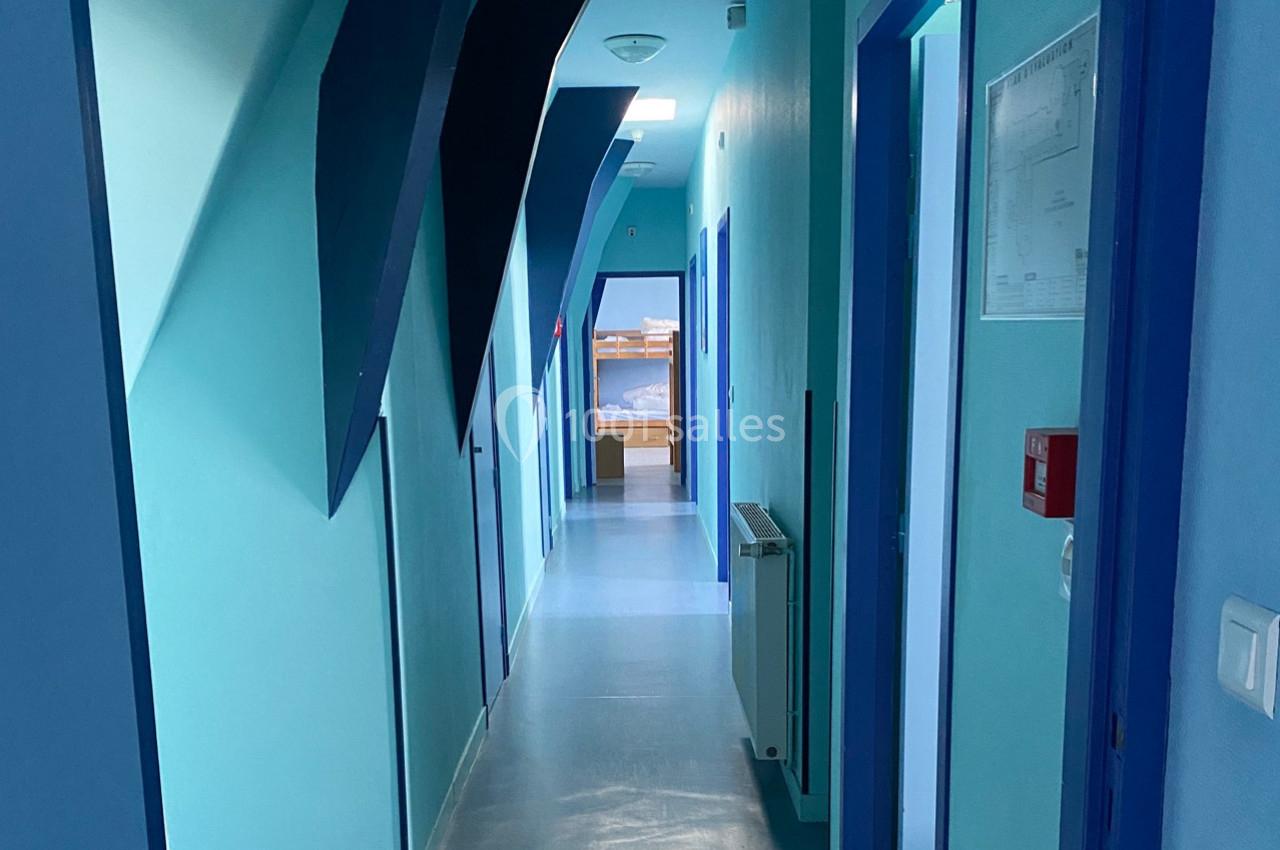 Couloir intérieur avec murs bleus, portes alignées des deux côtés et lumière naturelle au fond.