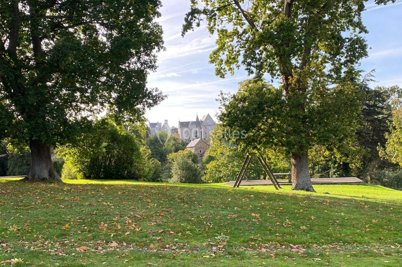Pelouse parsemée de feuilles d'automne, entourée d'arbres, avec un bâtiment en pierre visible à l'arrière-plan.