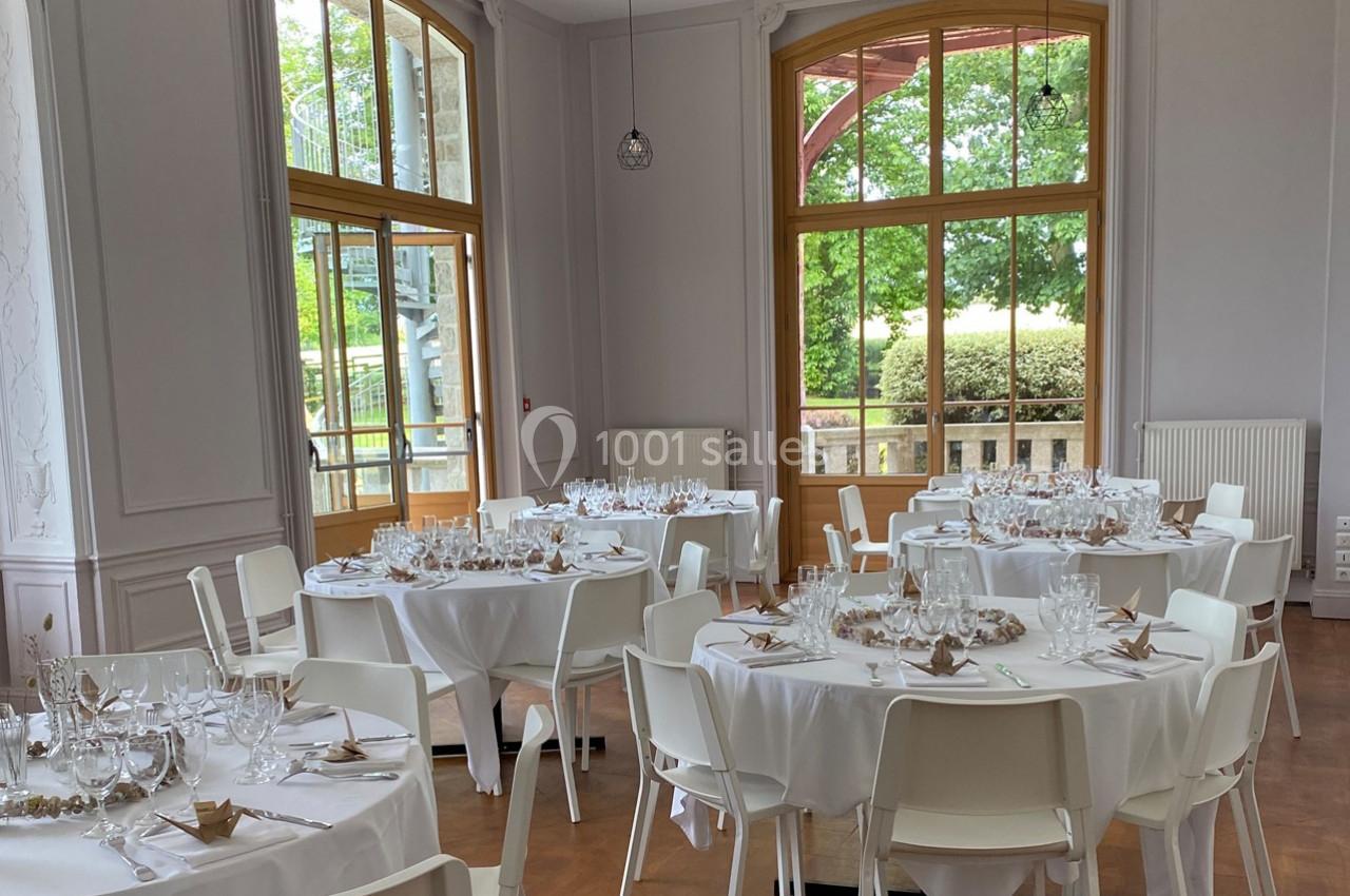 Salle lumineuse avec des tables rondes dressées pour un repas, entourées de chaises blanches, et grandes fenêtres donnant…