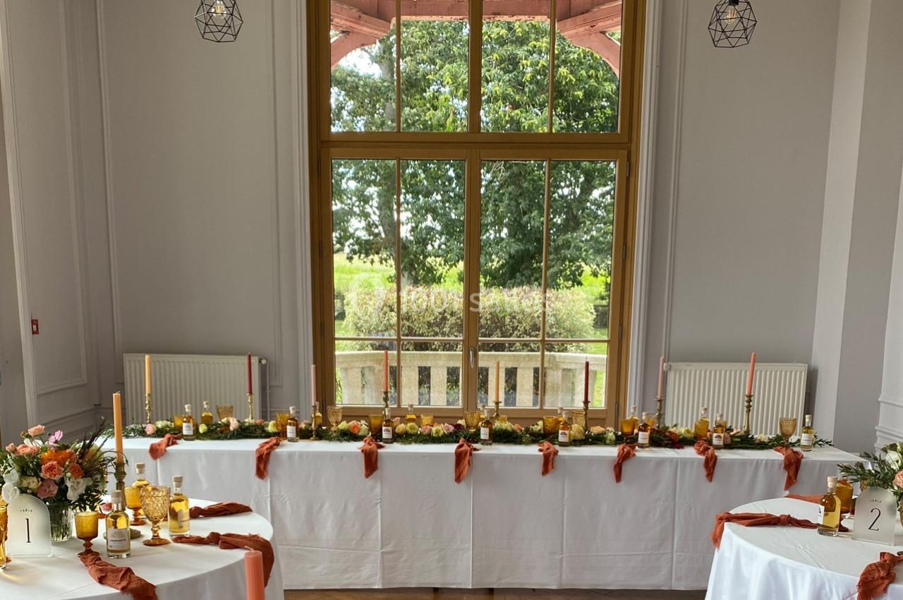 Salle décorée pour un événement avec des tables blanches ornées de bougies, fleurs et nappes orange, devant une grande…