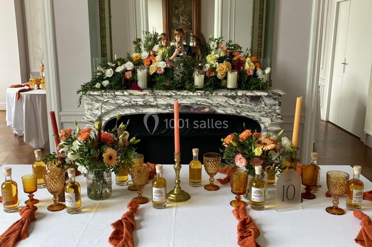 Table décorée avec des bougies, fleurs colorées et bouteilles, devant une cheminée ornée d'un miroir et d'arrangements…