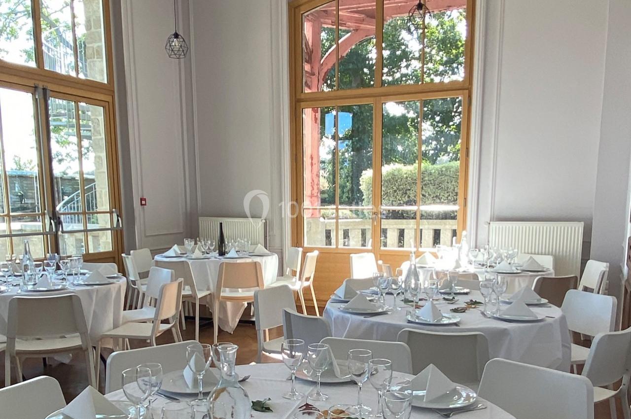 Salle lumineuse avec de grandes fenêtres, tables rondes dressées avec nappes blanches, verres et serviettes.