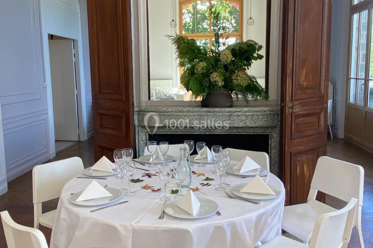 Table ronde dressée avec vaisselle et serviettes blanches, décorée de feuilles, dans une pièce lumineuse avec cheminée et…
