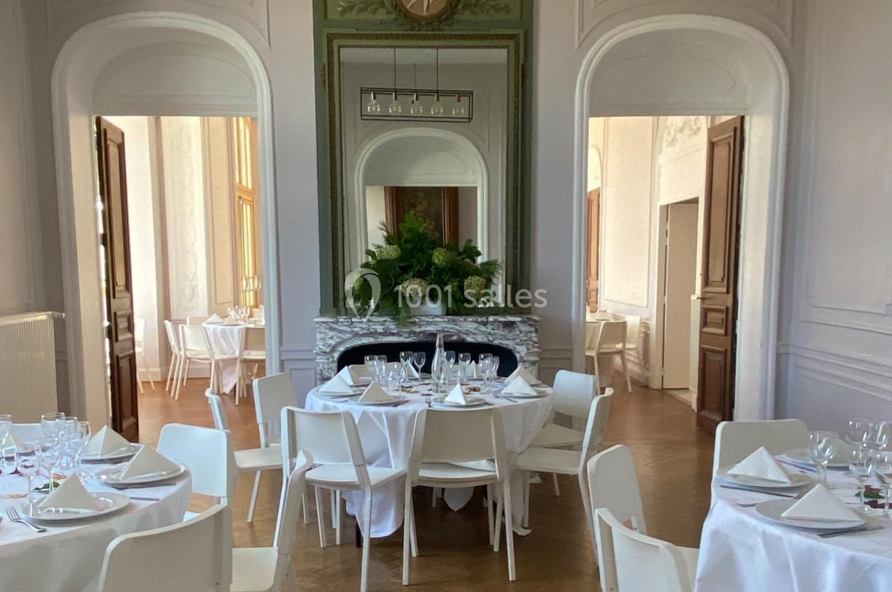 Salle élégante avec tables rondes dressées, chaises blanches et cheminée ornée de fleurs.