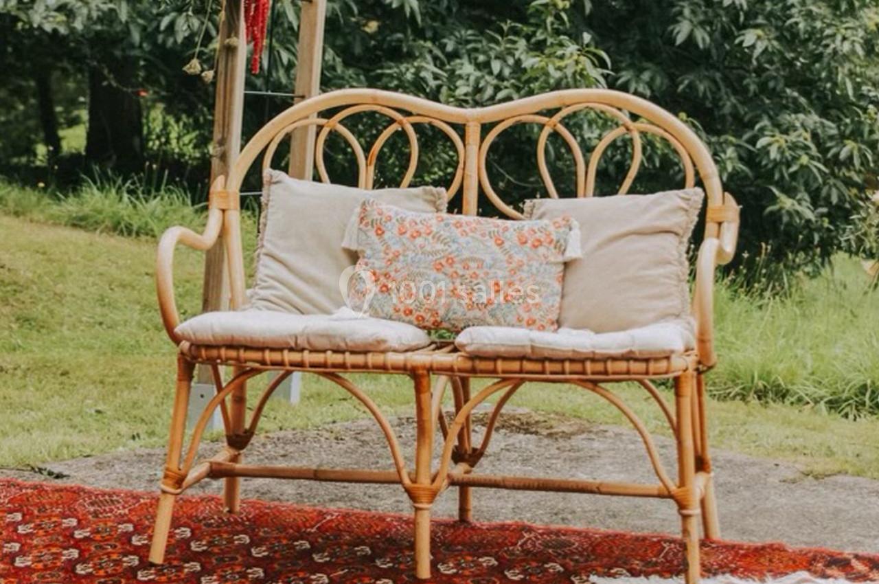 Banc en rotin avec coussins décoratifs, posé sur un tapis rouge dans un jardin verdoyant.