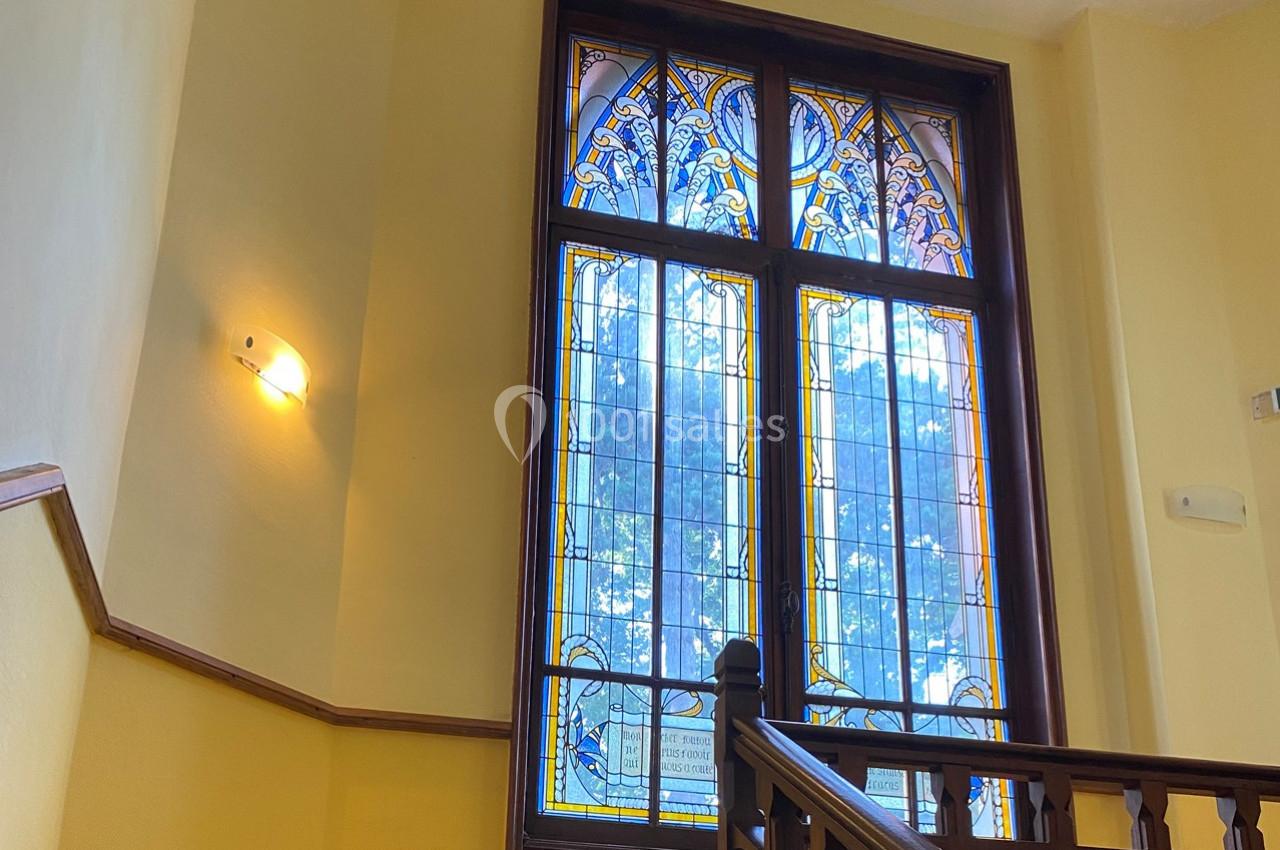 Fenêtre en vitrail coloré avec motifs décoratifs, encadrée de bois, éclairant un escalier intérieur.