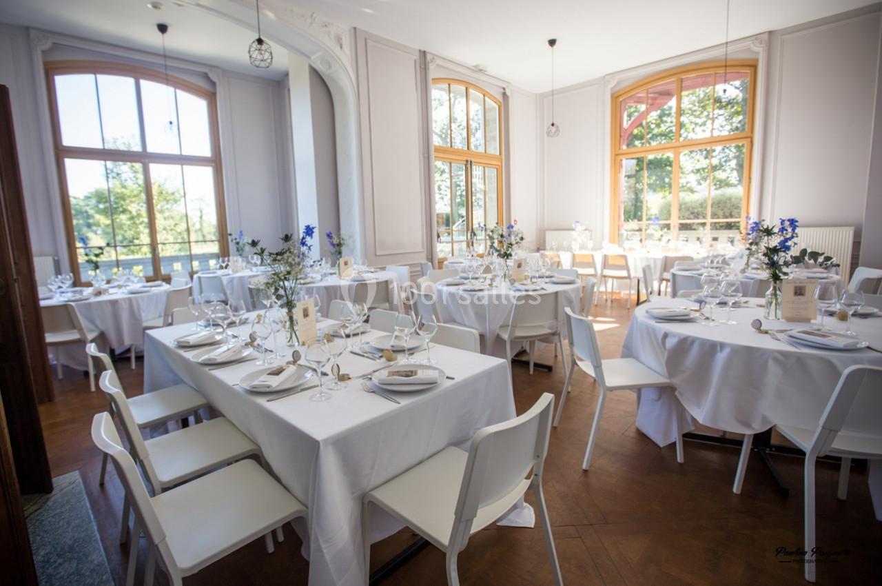 Salle lumineuse avec de grandes fenêtres, tables rondes et rectangulaires dressées pour un repas élégant.