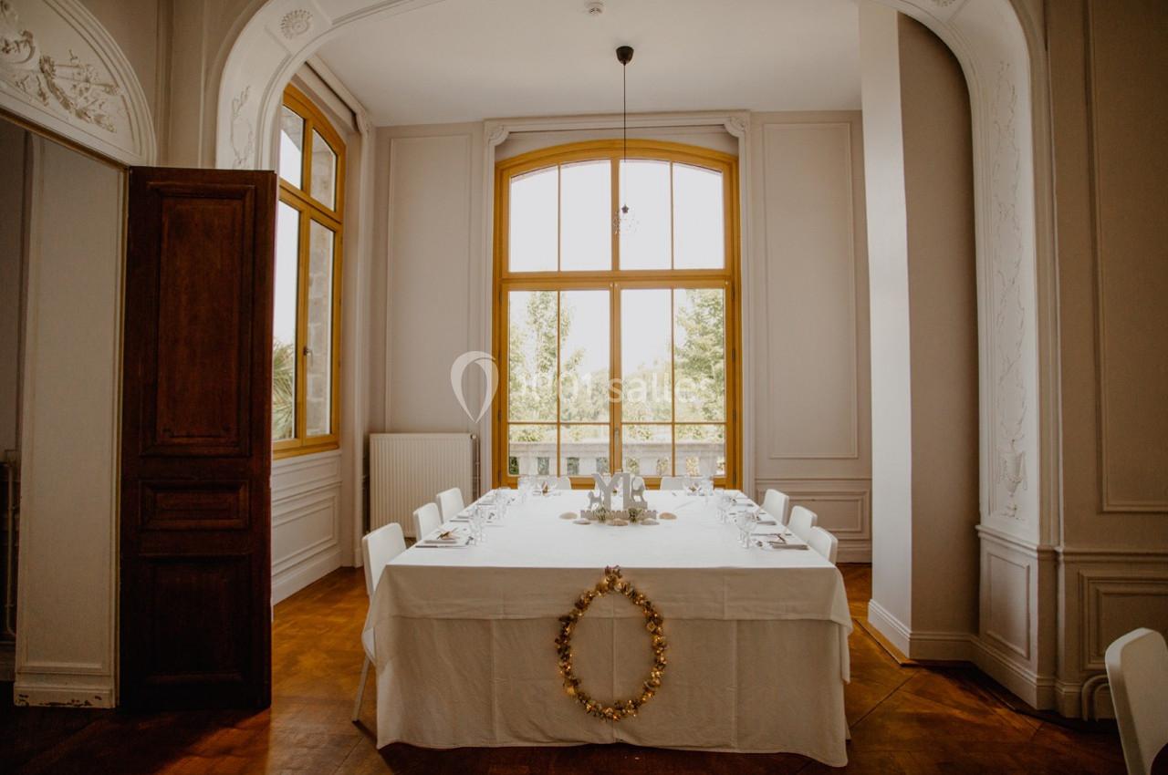 Salle à manger lumineuse avec une grande table dressée, entourée de chaises blanches, devant une fenêtre cintrée.