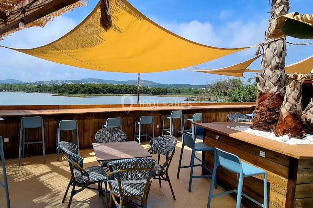 Terrasse en bois avec tables, chaises et voiles d'ombrage, offrant une vue sur un lac et des collines verdoyantes.