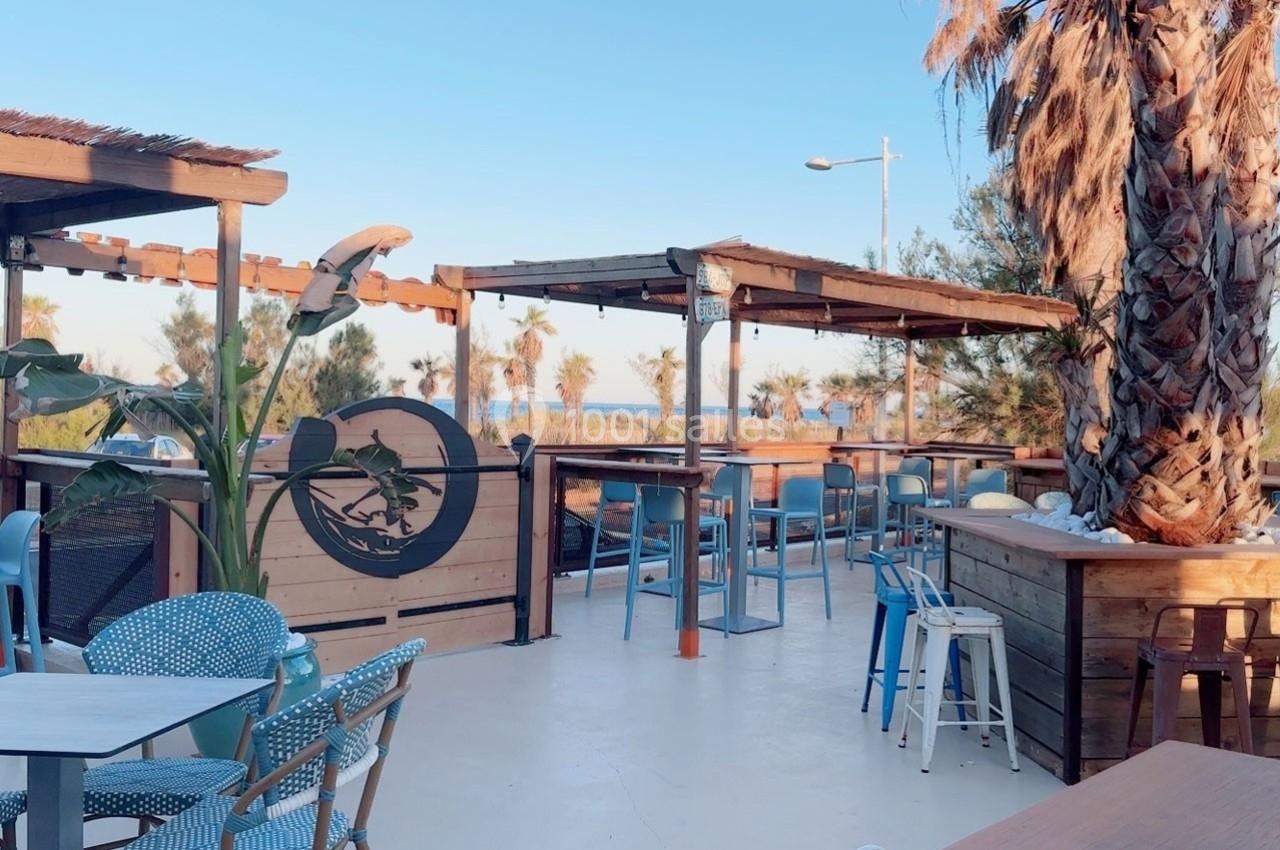Terrasse extérieure en bois avec tables et chaises, vue sur des palmiers et la mer sous un ciel dégagé.