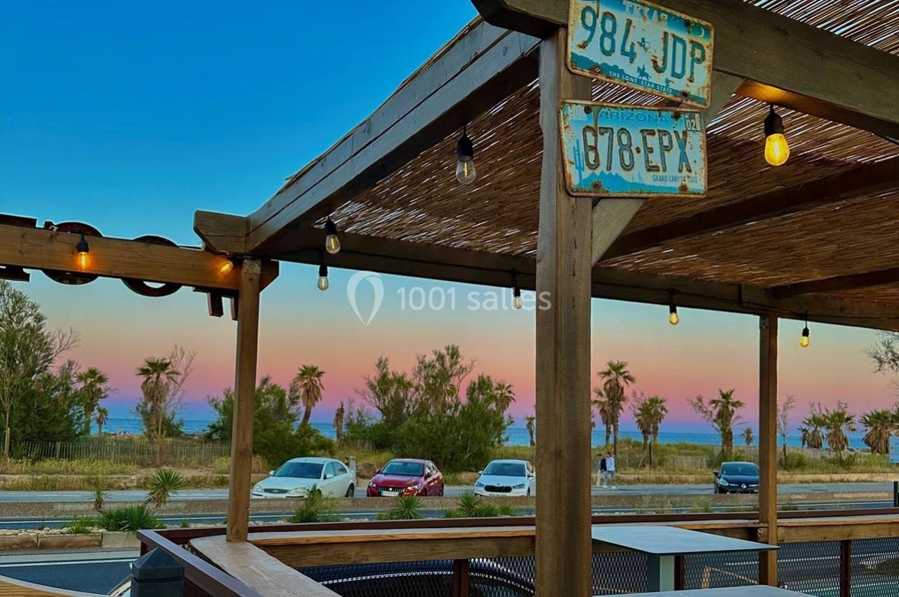 Terrasse en bois avec vue sur une route, des palmiers et un ciel coloré au coucher du soleil.