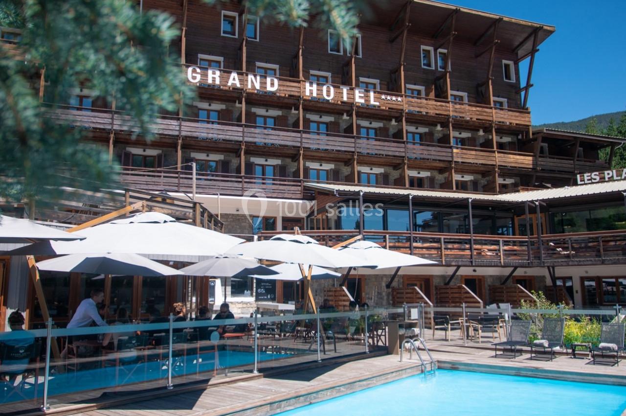 Façade d'un hôtel en bois avec terrasse, parasols et piscine extérieure sous un ciel bleu.