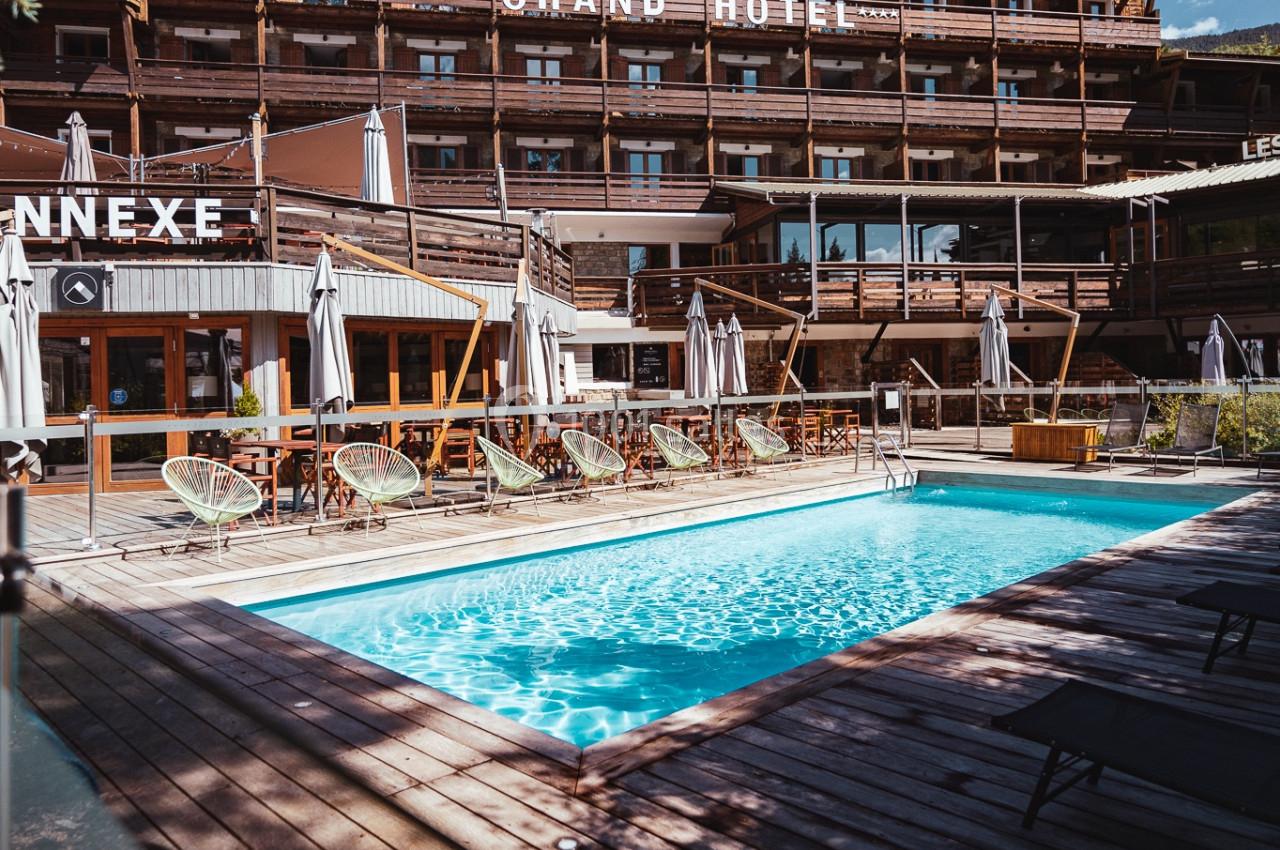 Piscine extérieure entourée de chaises longues et parasols, devant un grand bâtiment en bois sous un ciel dégagé.