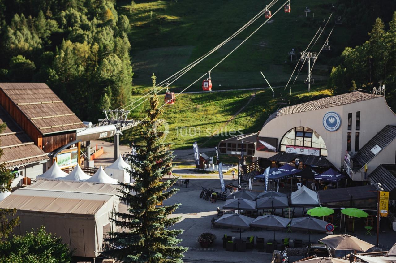 Vue d'une station de montagne avec piscine, terrasse, télésiège et forêt en arrière-plan par une journée ensoleillée.
