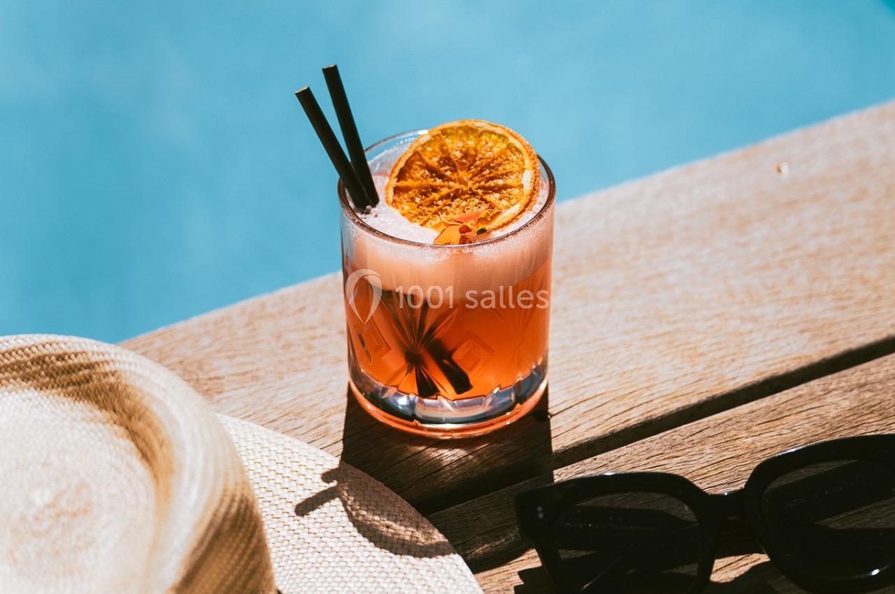 Verre de cocktail avec tranche d'orange, posé sur une table en bois près d'une piscine, avec un chapeau et des lunettes de…