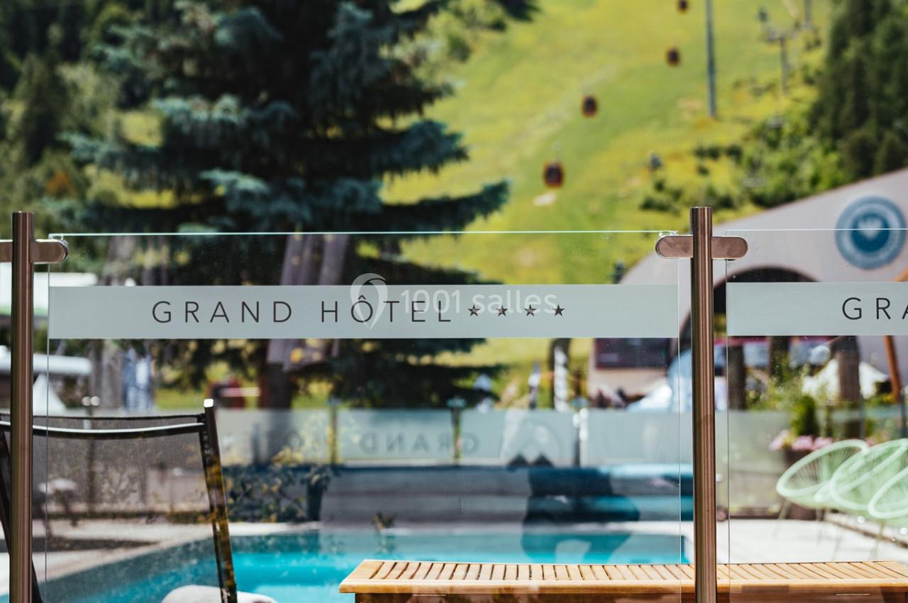 Vue sur une terrasse avec piscine, transat et panneau en verre marqué ’Grand Hôtel’, entourée de montagnes verdoyantes.