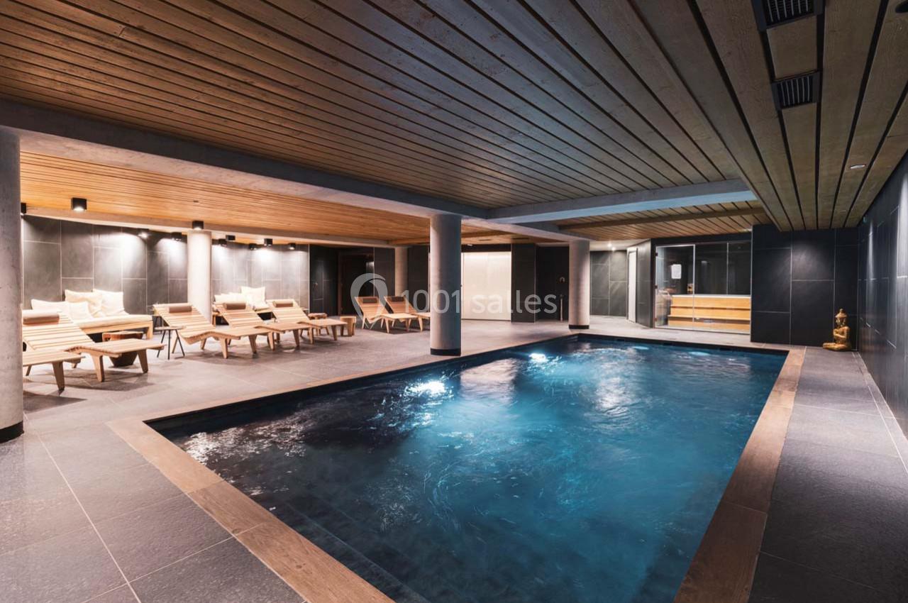 Piscine intérieure avec eau éclairée, entourée de chaises longues en bois et d'un plafond lambrissé.