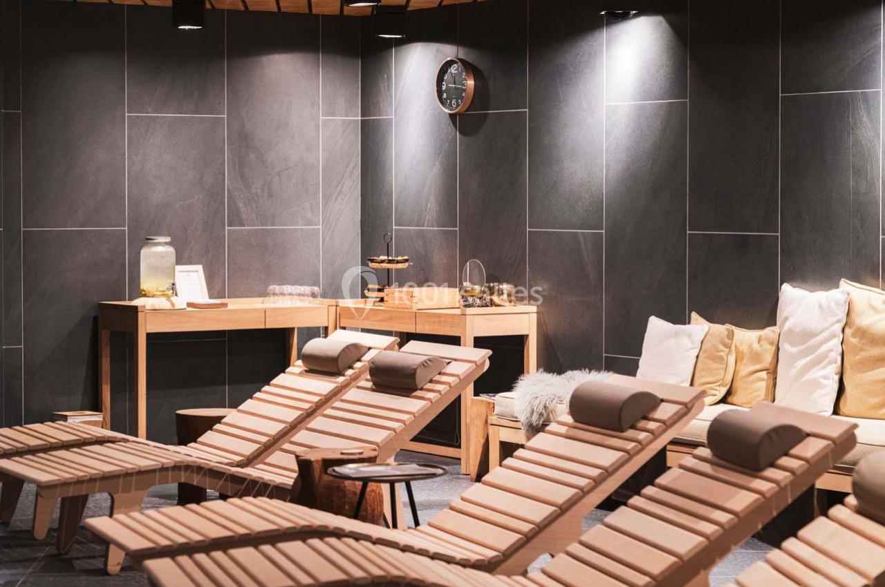 Chaises longues en bois alignées dans un espace détente avec murs sombres, coussins et table de rafraîchissements.