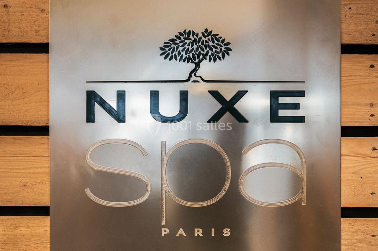 Plaque métallique avec le logo ’Nuxe Spa Paris’ gravé, fixée sur un mur en bois.