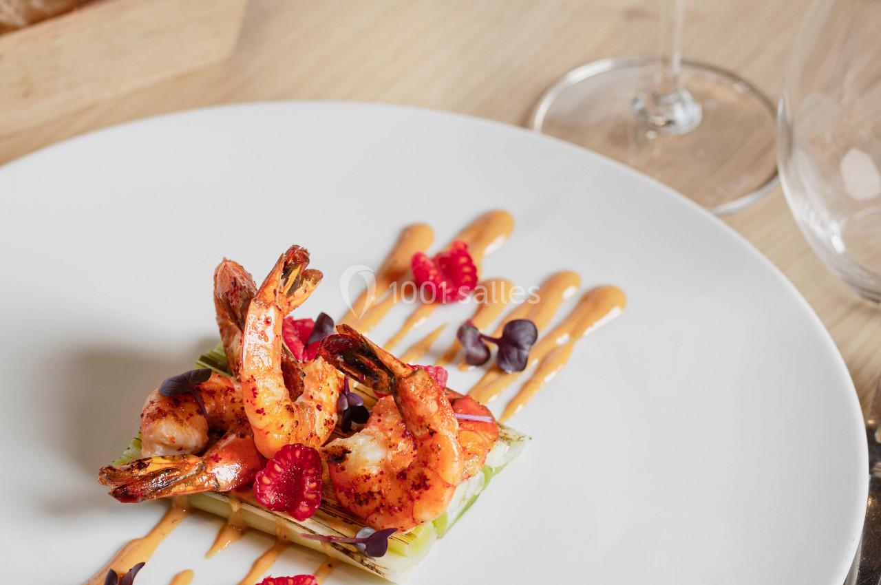 Assiette de crevettes grillées sur lit de légumes, décorée de framboises et sauce, avec verre de vin en arrière-plan.