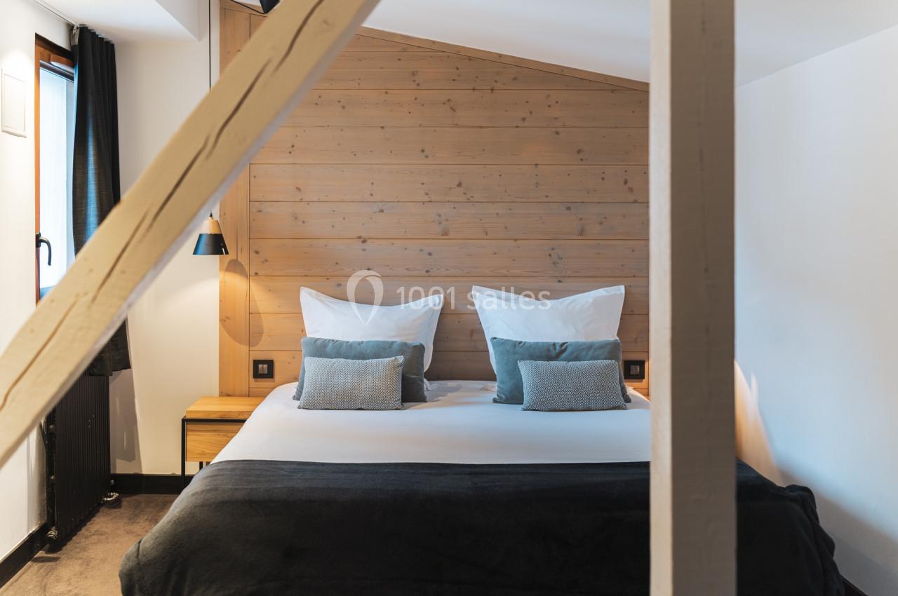 Lit double avec coussins et couverture sombre dans une chambre lumineuse aux murs en bois et poutres apparentes.
